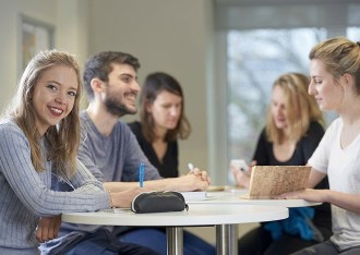 Groupe d’étudiants travaillant autour d’une table blanche dans une salle lumineuse, dont une étudiante souriante regardant vers l’objectif