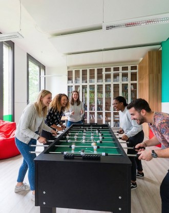 Étudiants jouant au baby-foot dans une salle de détente lumineuse avec bibliothèque en arrière-plan, au campus de Munich