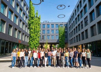 Groupe d’étudiants posant dans la cour moderne d’un campus à Munich, entourée de bâtiments gris et de colonnes végétalisées