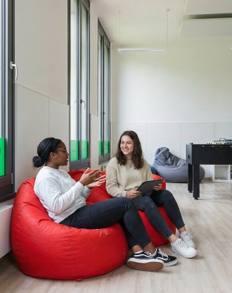 Deux étudiantes assises sur des poufs rouges discutent dans une salle de détente lumineuse du campus de Munich, avec un baby-foot en arrière-plan